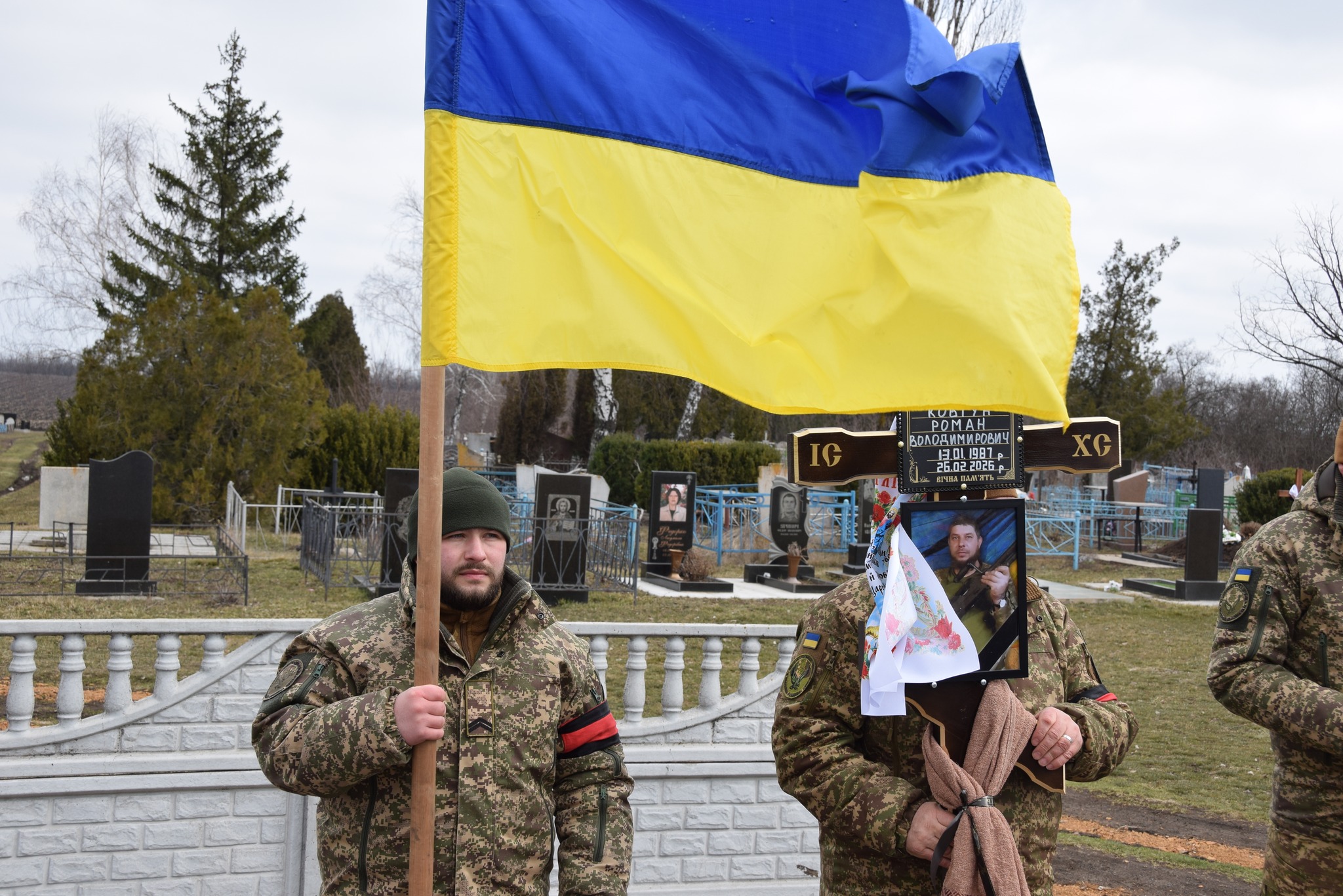 Воїни тримають прапор та намогильний хрест загиблого Романа Ковтуна